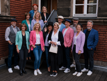 Copyright: Hans-Georg Leinberger / KI generiert Gruppe von Personen in bunter Kleidung und teils mit Hüten posiert lächelnd auf einer Treppe vor einem Backsteingebäude.