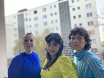 Drei Personen in blauem, gelbem und hellblauem Oberteil stehen nebeneinander vor einem hohen Wohnblock.