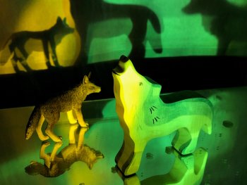 Zwei Wolf-Figuren, eine aus Holz mit heulender Kopfhaltung und eine detailreiche aus Kunststoff, werfen bei grünem und gelbem Licht große Schatten an die Wand.