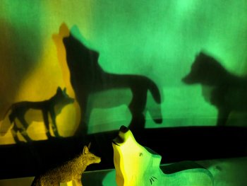 Foto: Kathrin Blüchert
Zwei Wolf-Figuren, eine aus Holz mit heulender Kopfhaltung und eine detailreiche aus Kunststoff, werfen bei grünem und gelbem Licht große Schatten an die Wand.