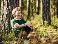 Eine lächelnde Person lehnt an einem Baumstamm im Wald und blickt entspannt nach oben in die Baumkronen.