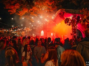 Menschenmenge feiert bei einem nächtlichen Open-Air-Konzert unter Bäumen, während die Bühne in rotes und oranges Licht getaucht ist.