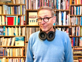 Person mit Brille und Kopfhörern um den Hals steht lächelnd vor einem großen Bücherregal.