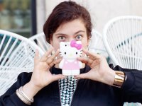 Eine Person hält sich eine Handyhülle in Form einer Hello-Kitty-Figur vor das Gesicht und blickt über den Rand hinweg in die Kamera.