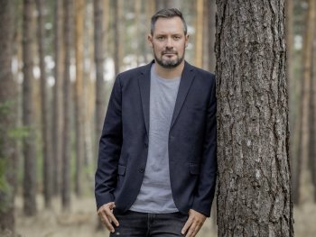 Copyright: Peter von Felbert Person mit dunklem Sakko und grauem T-Shirt lehnt an einem Baumstamm in einem Kiefernwald.