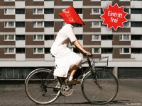 Person in weißem Kleid mit rotem Tuch auf dem Kopf fährt Fahrrad vor einem mehrstöckigen Wohnhaus