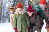 Zwei Kinder und zwei Erwachsene in warmer Winterkleidung mit bunten Mützen betrachten gemeinsam etwas am Boden in einem verschneiten Wald.