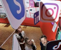 Drei Personen blicken in einer Ausstellung nach oben zwischen großen Icons bekannter Social-Media-Plattformen wie Instagram, LinkedIn und Skype.