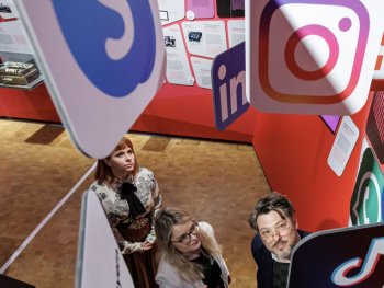 Drei Personen blicken in einer Ausstellung nach oben zwischen großen Icons bekannter Social-Media-Plattformen wie Instagram, LinkedIn und Skype.