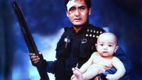Verletzter Mann hält eine Schrotflinte in einer Hand und ein Baby im anderen Arm.