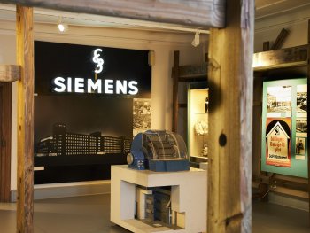 Museumsausstellung mit beleuchtetem Siemens-Firmenlogo über einem historischen Foto eines Fabrikgebäudes und einem technischen Ausstellungsobjekt auf einem Sockel.
