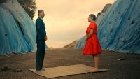 Ein Mann im Anzug und eine Frau im roten Kleid stehen sich auf einer Matte gegenüber, umgeben von blau verhüllten Hügeln.