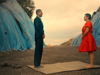 Copyright: Neufilm
Ein Mann im Anzug und eine Frau im roten Kleid stehen sich auf einer Matte gegenüber, umgeben von blau verhüllten Hügeln.