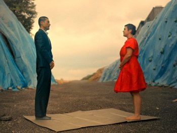 Copyright: Neufilm Ein Mann im Anzug und eine Frau im roten Kleid stehen sich auf einer Matte gegenüber, umgeben von blau verhüllten Hügeln.
