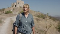 Eine Frau mit Rucksack wandert auf einem Schotterweg durch eine trockene Hügellandschaft, im Hintergrund steht eine alte Ruine auf einem Hügel.