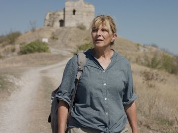 Copyright: Piffl Medien, Komplizen Film
Eine Frau mit Rucksack wandert auf einem Schotterweg durch eine trockene Hügellandschaft, im Hintergrund steht eine alte Ruine auf einem Hügel.