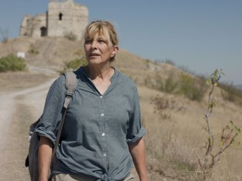 Copyright: Piffl Medien, Komplizen Film Eine Frau mit Rucksack wandert auf einem Schotterweg durch eine trockene Hügellandschaft, im Hintergrund steht eine alte Ruine auf einem Hügel.