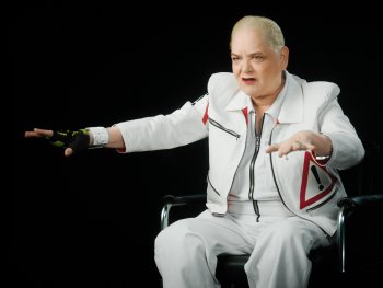 Copyright: BASIS BERLIN Filmproduktion; ZDF; Thomas Lütz
Hella von Sinnen mit weißem Anzug und Warnsymbol auf der Jacke sitzt gestikulierend auf einem Stuhl vor schwarzem Hintergrund.