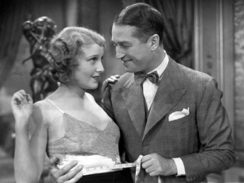 Foto: Images courtesy of Park Circus; Universal
Schwarz-Weiß-Filmszene, in der ein Mann im Anzug mit einem Maßband die Taille einer Frau in einem Spitzenkleid misst, während sich beide anlächeln.