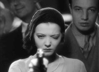 Schwarz-weißes Filmstandbild einer jungen Frau mit Mütze, die konzentriert mit einer Pistole in die Kamera blickt.
