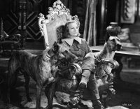 Schwarz-Weiß-Filmszene einer Person in historischem Kostüm auf einem verzierten Thronsessel, flankiert von zwei großen Doggen in einem prunkvollen Saal.