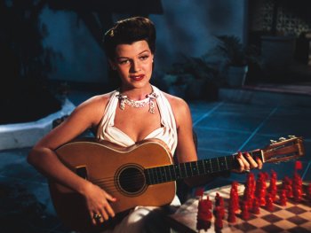 Copyright: Images courtesy of Park Circus; Universal
Frau im weißen Kleid mit Diamantcollier spielt Gitarre neben einem Schachbrett mit roten Figuren.