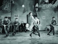 Schwarz-weiße Filmszene mit einer tanzenden Frau im Kleid inmitten mehrerer Männer, die in einem alten Saal Steppschritte üben.