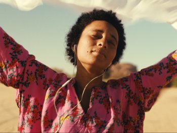 Copyright: Pili Films Person in pinkfarbener geblümter Bluse hält mit erhobenen Armen ein weißes Tuch in den Wind, mit Kopfhörern und geschlossenen Augen in einer sandigen Landschaft.