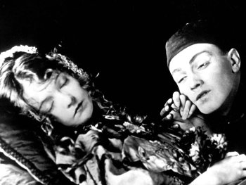 Copyright: Filmverleih Schwarz-Weiß-Filmszene, in der eine Person mit geschlossenen Augen ruht, während eine zweite Person mit Kopfbedeckung sich ihr nah zuneigt.