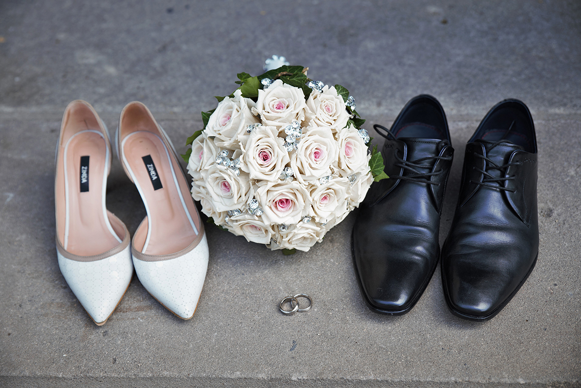 Weiße Damenschuhe und schwarze Herrenschuhe stehen neben einem Brautstrauß aus weißen und rosa Rosen sowie zwei Eheringen auf einer Steinfläche.