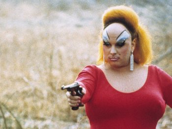 Copyright: Filmverleih
Person mit auffälligem Make-up, hochtoupierten roten Haaren und rotem Kleid zielt mit einer Pistole in einer trockenen Landschaft.