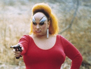 Copyright: Filmverleih Person mit auffälligem Make-up, hochtoupierten roten Haaren und rotem Kleid zielt mit einer Pistole in einer trockenen Landschaft.
