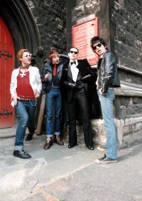 Vier Personen in Punk- und Rockabilly-Kleidung mit Sonnenbrillen und Lederjacken posieren vor dem Eingang einer Kirche.