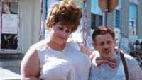 Zwei Personen in auffälliger Retro-Kleidung stehen eng nebeneinander auf einer Straße, im Hintergrund ein Schild mit der Aufschrift „We love Tracy“.