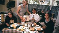 Eine vierköpfige Familie sitzt lächelnd am reich gedeckten Frühstückstisch, während die Mutter eine Cornflakes-Packung hält.