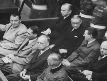 Copyright: Bundesarchiv Abteilung audiovisuelle Medien
Historische Schwarz-Weiß-Aufnahme zeigt die Angeklagten der Nürnberger Prozesse in Anzügen auf einer Bank sitzend, bewacht von einem Soldaten mit Helm.