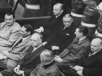 Copyright: Bundesarchiv Abteilung audiovisuelle Medien Historische Schwarz-Weiß-Aufnahme zeigt die Angeklagten der Nürnberger Prozesse in Anzügen auf einer Bank sitzend, bewacht von einem Soldaten mit Helm.