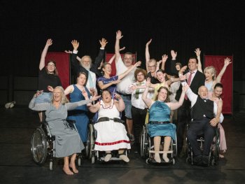Foto: Sebastian Autenrieth
Eine große Gruppe von Menschen posiert fröhlich mit erhobenen Armen auf einer Theaterbühne.