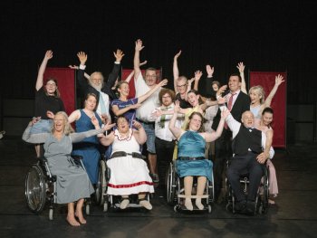 Eine große Gruppe von Menschen posiert fröhlich mit erhobenen Armen auf einer Theaterbühne.