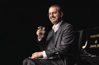 Lächelnder Mann im Anzug mit Headset-Mikrofon sitzt neben einem Flügel und hält ein Wasserglas in der Hand.