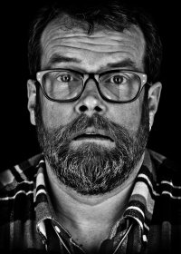 Schwarz-weißes Porträt einer bärtigen Person mit Brille und Karohemd, die mit ernstem Blick direkt in die Kamera schaut.