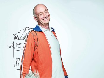 Copyright: Guido Werner Lächelnder Mann in orangefarbener Trainingsjacke trägt einen gezeichneten Schulrucksack mit Stiften und Lineal auf der Schulter.