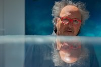 Ein Mann mit roter Brille und wirrem Haar blickt über eine spiegelnde Fläche, in der sich sein Gesicht reflektiert.