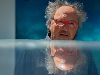 Copyright: Michael Palm Ein Mann mit roter Brille und wirrem Haar blickt über eine spiegelnde Fläche, in der sich sein Gesicht reflektiert.