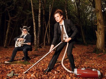 Copyright: Carsten Bockermann Eine Person in Anzug saugt mit einem altmodischen Staubsauger Laub im Wald, während im Hintergrund eine Person mit Hörnerhelm und Gitarre auf einem Baumstumpf sitzt.