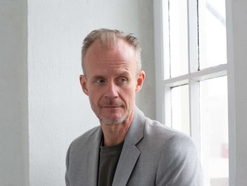Copyright: Roland Schmid Porträt eines Mannes mit grauem Blazer, der neben einem Fenster steht und zur Seite blickt.