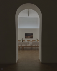 Blick durch einen Rundbogen in einen hellen Saal mit langer, festlich gedeckter Tafel und Blumenschmuck vor einer Wand mit gerahmtem Bild.