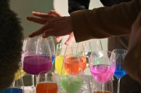 Nahaufnahme mehrerer Weingläser, die zur Hälfte mit bunter Flüssigkeit in unterschiedlichen Farben gefüllt ist. Es ist eine Hand zu sehen, die mit einem Finger den Glasrand berührt, um einen Ton zu erzeugen.