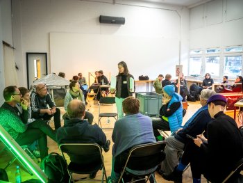 Foto: Christian Bartsch
Eine Person steht mit Ordnern in der Hand vor einer im Stuhlkreis sitzenden Gruppe in einem hellen Raum mit weiteren Sitzgruppen im Hintergrund.