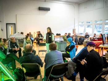 Eine Person steht mit Ordnern in der Hand vor einer im Stuhlkreis sitzenden Gruppe in einem hellen Raum mit weiteren Sitzgruppen im Hintergrund.
